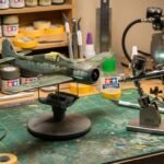 Bancada de plastimodelismo com modelo de avião militar em escala sendo pintado com aerógrafo de dupla ação Iwata sobre base de corte.